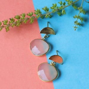 Può includere: Un paio di orecchini color oro con una parte superiore a mezzaluna e una grande gemma ovale rosa. Le gemme hanno un bordo dorato e sono sospese dalla forma a mezzaluna. Su sfondo rosa e blu.