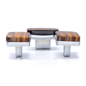 May include: Three cabinet knobs with a rectangular, tiger-eye-patterned top and a silver-colored base. The knobs are arranged in a row, with the center knob slightly larger than the two flanking knobs. The background is white.