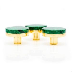 Puede incluir: Tres tiradores decorativos para armarios con una parte superior redonda de color verde malaquita y una base dorada. Los tiradores están dispuestos en fila sobre un fondo blanco. La parte superior verde tiene un diseño veteado.