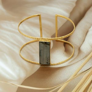 Könnte beinhalten: Ein goldfarbenes Armreif-Armband mit einem rechteckigen, grauen Stein-Mittelstück. Das Armband hat eine strukturierte Oberfläche und ein Doppelband-Design mit vertikalen Stäben, die die Bänder verbinden.