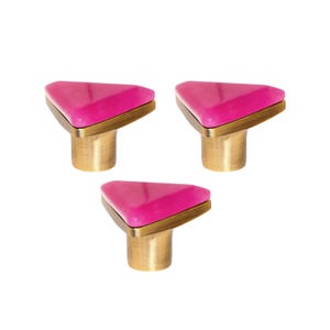 May include: Three cabinet knobs with a triangular, hot pink top and a brass base. The knobs have a modern design and are set against a white background. The knobs are likely for furniture or home decor.
