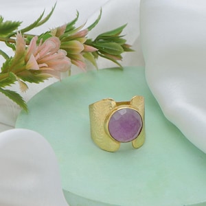 Pode incluir: Um anel dourado com uma grande pedra preciosa redonda de cor roxa clara. O anel tem uma banda texturizada com um padrão floral. A pedra preciosa é definida em uma configuração de moldura. O anel é exibido em uma superfície verde clara.