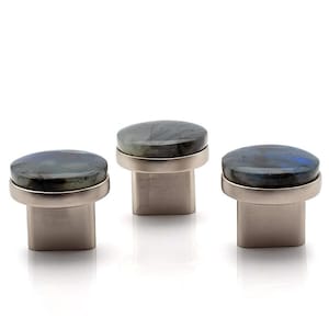 Op de afbeelding: Drie kastknoppen met een ronde, gepolijste stenen bovenkant in tinten blauw, groen en grijs. De steen is gezet in een zilverkleurige metalen ring en gemonteerd op een vierkante basis. Deze knoppen zijn ideaal om een vleugje elegantie aan meubels toe te voegen.