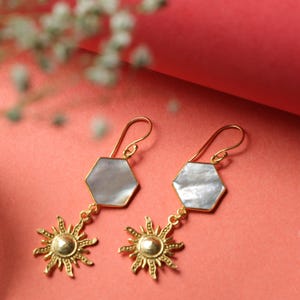 Puede incluir: Un par de pendientes dorados con diseño de sol y hexágono. Los pendientes tienen ganchos dorados y una piedra nacarada en forma de hexágono. Debajo del hexágono cuelga un colgante de sol detallado. El fondo es de color coral.