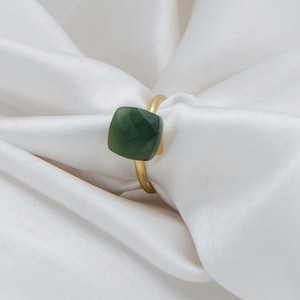 May include: A gold ring featuring a square, faceted, dark green gemstone. The ring is set on a white satin background, highlighting the gold band and the gemstone's color and shape. The ring is a piece of jewelry.