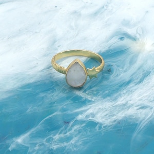 May include: A gold-colored ring with a pear-shaped, white gemstone. The ring has a textured band and a small star detail on the side. The ring is set against a blue and white background.