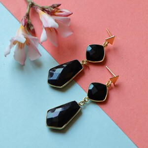 May include: Black gemstone dangle earrings with gold-colored trim and posts. The earrings feature a square top stone and a larger, faceted bottom stone. A sprig of pink and white flowers is in the background.