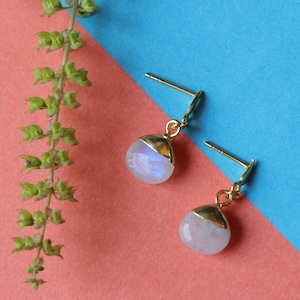May include: A pair of gold-coloured stud earrings featuring round, iridescent moonstone drops. The earrings have a gold cap, set against a pink and blue background. These earrings are a type of jewellery.
