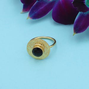 Könnte beinhalten: Ein goldfarbener Ring mit einer kreisförmigen, gravierten Oberfläche und einem schwarzen Edelstein in der Mitte. Der Ring befindet sich vor einem hellblauen Hintergrund, mit violetten Orchideenblütenblättern in der oberen rechten Ecke. Das Ringdesign hat ein Blumenmuster.