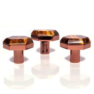 May include: Three cabinet knobs with a geometric design. Each knob features a faceted, amber-coloured gem set atop a copper-coloured, cylindrical base. The knobs are arranged against a white background, with their reflections visible.