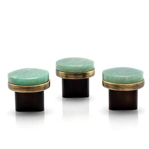 Könnte beinhalten: Drei dekorative Schrankknöpfe. Jeder Knopf hat eine runde, hellgrüne Steinplatte, ein messingfarbenes Band und einen dunkelbraunen, quadratischen Sockel. Die Knöpfe sind vor einem weißen Hintergrund angeordnet.