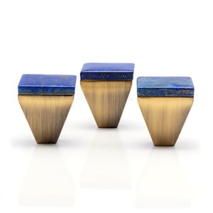 Peut inclure: Trois boutons de meuble avec une finition en laiton et un dessus carré en pierre bleu foncé. Les boutons ont une forme triangulaire et sont posés sur un fond blanc. Ces boutons décoratifs ajoutent une touche d'élégance à tout meuble.