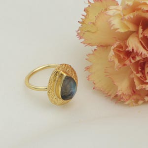 May include: A gold ring featuring an oval, textured setting and a blue-grey gemstone. The ring is set against a white background, with a soft yellow and pink flower in the background.