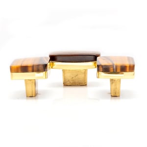 May include: A set of three cabinet knobs. Each knob has a square, amber-coloured top with a gold-coloured base. The centre knob is a darker brown colour. The knobs are designed for furniture and home decor.