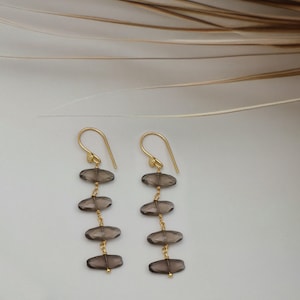 May include: A pair of gold-coloured drop earrings. Each earring has a hook and chain design, with four oval, smoky-grey gemstones. The earrings are presented on a white surface with a neutral background.