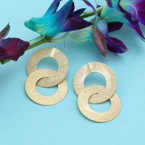 May include: A pair of gold-coloured, textured metal earrings featuring an interlocking circle design. The earrings are displayed against a light blue backdrop, with a vibrant purple and teal flower in the upper part. The earrings have a matte finish.