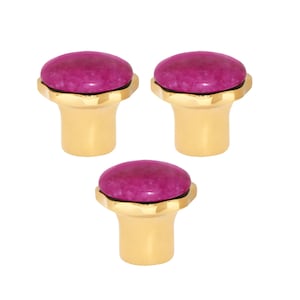 May include: Three decorative cabinet knobs. Each knob features a round, magenta-coloured stone set atop a gold-coloured base. The base has a fluted design around the stone and a cylindrical stem.