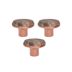 以下が含まれることがあります： 3つの装飾的なキャビネットノブ。各ノブは、銅色のベースと、ピンク、緑、茶色のマーブル模様のファセットトップが特徴です。ノブは、家具またはキャビネットのハードウェア用に設計されています。