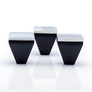 May include: Three black, geometric containers with white, rectangular tops. The containers have a trapezoidal shape, with a wider top and a narrower base. The white tops appear to be made of a translucent material, and the containers are set against a white background.
