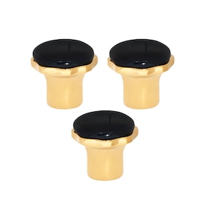 May include: Three cabinet knobs with a gold-coloured base and a black, oval-shaped top. The knobs have a decorative, scalloped edge where the black top meets the gold base. The knobs are arranged in a triangular pattern.