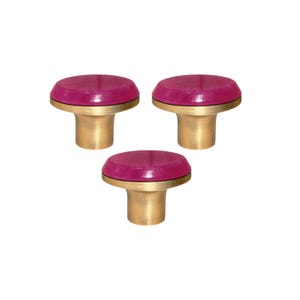 Può includere: Tre pomelli decorativi per mobili. Ogni pomello presenta una parte superiore rotonda color magenta con una sottile fascia nera, posta su una base cilindrica dorata. I pomelli sono disposti su uno sfondo bianco.