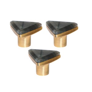 Puede incluir: Tres tiradores de armario con tapas triangulares de piedra gris y bases de color latón. Los tiradores tienen un diseño moderno y están sobre un fondo blanco. Cada tirador tiene un borde dorado.