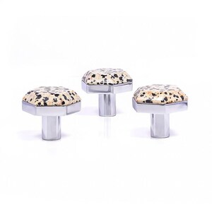 Op de afbeelding: Drie decoratieve kastknoppen met een gespikkelde, crèmekleurige bovenkant en een zilverkleurige, geometrische basis. De knoppen zijn geplaatst tegen een witte achtergrond, wat hun ontwerp en potentiële gebruik in huisdecoratie benadrukt.