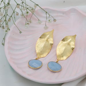 May include: A pair of gold-colored leaf-shaped earrings with a textured finish. Each earring features a light blue, oval-shaped stone with a gold-colored border. The earrings are displayed on a pink, marbled surface.