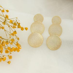 May include: Gold-coloured, textured disc earrings. Each earring has three graduated circles, creating a cascading effect. The earrings are displayed on a white, textured surface, with yellow floral accents in the background. A simple, elegant jewellery design.