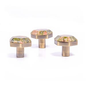 Può includere: Tre pomelli decorativi per mobili con un design geometrico. Ogni pomello presenta una parte superiore in pietra sfaccettata multicolore nei toni del verde, rosa e marrone, montata su una base color ottone. I pomelli sono esposti su sfondo bianco.