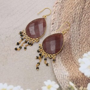 May include: A pair of gold-colored earrings with teardrop-shaped, faceted, reddish-brown stones. The earrings have a fringe of small black beads and gold-colored accents. The earrings are designed with a hook closure.