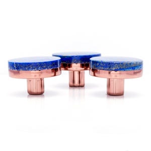 May include: Three cabinet knobs with a copper base and a blue lapis lazuli top. The knobs are round and have a polished finish. The copper base is cylindrical. The lapis lazuli top has a speckled appearance. The knobs are suitable for cabinets.