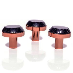 Könnte beinhalten: Drei dekorative Schrankknöpfe. Jeder Knopf hat einen kupferfarbenen Sockel und Stiel, gekrönt von einer dunklen, gesprenkelten Kappe. Die Kappen haben einen metallischen Glanz. Die Knöpfe sind vor weißem Hintergrund angeordnet.