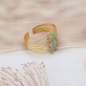 Puede incluir: Un brazalete de puño dorado con un diseño texturizado. El brazalete presenta múltiples bandas paralelas y está adornado con un grupo de piedras preciosas de color azul claro. El diseño abierto permite un uso fácil y se adapta a diferentes tamaños de muñeca.