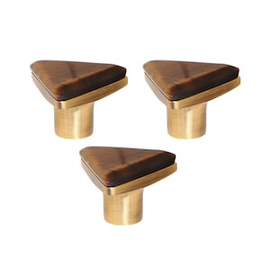 May include: Three cabinet knobs. Each has a triangular, brown, stone-like top and a brass-coloured base. The knobs have a modern design and are set against a white background. Suitable for drawers and cabinets.