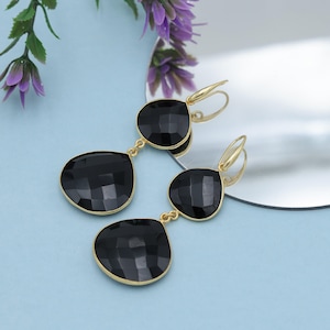 May include: Gold-coloured drop earrings featuring three faceted black teardrop-shaped stones. The earrings are presented on a light blue surface with a reflective surface and purple flowers. The earrings have a lever back closure.