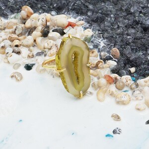May include: A gold ring with a large, oval agate stone. The agate is light green and cream with a lighter central band. The ring is set against a background of small pebbles and a white surface.