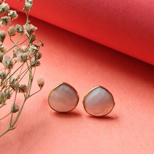 May include: Two pear-shaped stud earrings with a gold-coloured trim. The earrings have a light pinkish-white centre. The background is a gradient of pink and red, with a sprig of white flowers in the upper left corner.