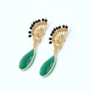 May include: A pair of gold-colored earrings with a peacock design. The earrings feature a fan-shaped top with black bead accents and a teardrop-shaped, faceted green stone pendant. The earrings are set against a white background.