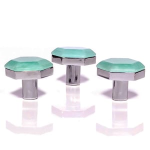 Può includere: Tre pomelli decorativi per mobili dal design geometrico. Ogni pomello presenta una parte superiore ottagonale verde chiaro e una base cilindrica argentata. I pomelli sono disposti in fila su sfondo bianco.