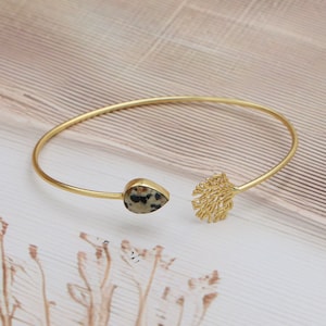 May include: A gold-coloured open cuff bracelet. One end features a teardrop-shaped stone with black and tan spots. The other end has a decorative, openwork tree design. The bracelet is displayed on a light-coloured, textured surface.