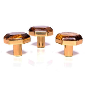 May include: Three cabinet knobs with an octagonal amber top and a gold-coloured base and stem. The knobs are displayed in a row. The amber tops have a patterned appearance. The knobs are suitable for cabinets or drawers.