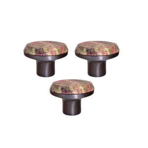 May include: A set of three cabinet knobs. Each knob has a round top with a floral pattern in pink, green, and brown. The base is a dark metallic colour. These knobs are suitable for furniture and home decor.