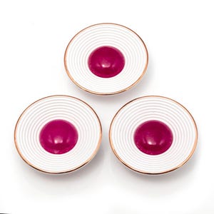 Puede incluir: Tres platos decorativos blancos y dorados con un diseño concéntrico acanalado. Cada plato presenta un acento central circular de color magenta intenso. Los platos están dispuestos en un patrón triangular sobre un fondo blanco.