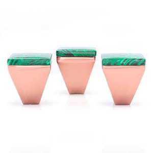 Può includere: Tre pomelli decorativi per mobili. Ogni pomello presenta una parte superiore quadrata in malachite verde e una base geometrica color rame. I pomelli sono disposti in fila su uno sfondo bianco, evidenziando il loro design e contrasto cromatico.