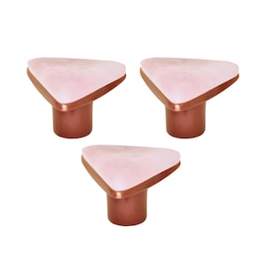 Pode incluir: Três puxadores decorativos para armários. Cada puxador apresenta um topo triangular rosa claro com efeito marmoreado branco, uma borda cor de cobre e uma base cilíndrica cor de cobre.