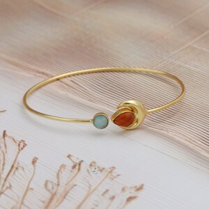 Może przedstawiać: Złota bransoletka z dwoma akcentami z kamieni szlachetnych. Jednym z nich jest okrągły, jasnoniebieski kamień, a drugim pomarańczowy kamień w kształcie łzy, osadzony w kształcie półksiężyca. Bransoletka leży na jasnej powierzchni.