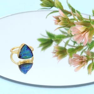 Könnte beinhalten: Ein goldener Ring mit einem dreieckigen, irisierenden blau-grünen Stein. Der Ring liegt auf einer reflektierenden Oberfläche, mit einem verschwommenen Blumenarrangement im Hintergrund. Das Ringdesign hat ein Doppelband.