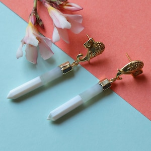 Könnte beinhalten: Ein Paar goldfarbene Ohrringe in Elefantenform mit weißen und hellgrauen Kristallanhängern. Die Ohrringe werden auf einem hellblauen und korallenfarbenen Hintergrund mit rosa Blüten in der oberen linken Ecke präsentiert.