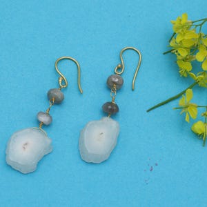 Puede incluir: Un par de pendientes dorados con un diseño único. Cada pendiente presenta un gancho dorado, cuentas de piedra grises y un colgante grande de piedra blanca y gris de forma irregular. Los pendientes se exhiben sobre un fondo azul.